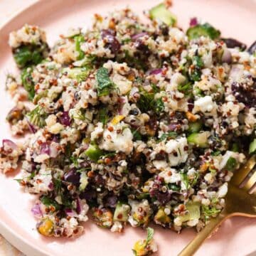 A white plate with a serving of quinoa salad mixed with diced cucumbers, chopped herbs, red onions, nuts, and feta cheese. A gold fork is resting on the plate. The background is a light, textured surface.
