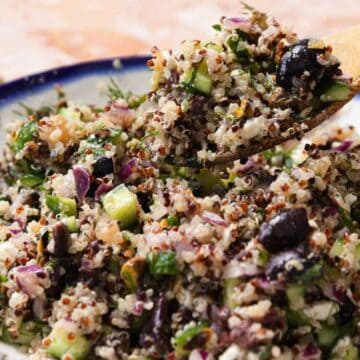 A bowl of quinoa salad is being mixed with a wooden spoon. The salad contains quinoa, chopped cucumbers, black olives, red onions, and herbs, all in a ceramic dish with a blue rim.