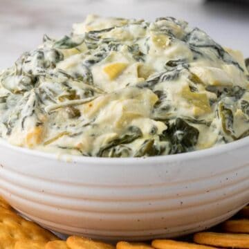 A white bowl filled with creamy spinach and artichoke dip, placed on a marble surface. The dip is surrounded by a layer of round, golden crackers.