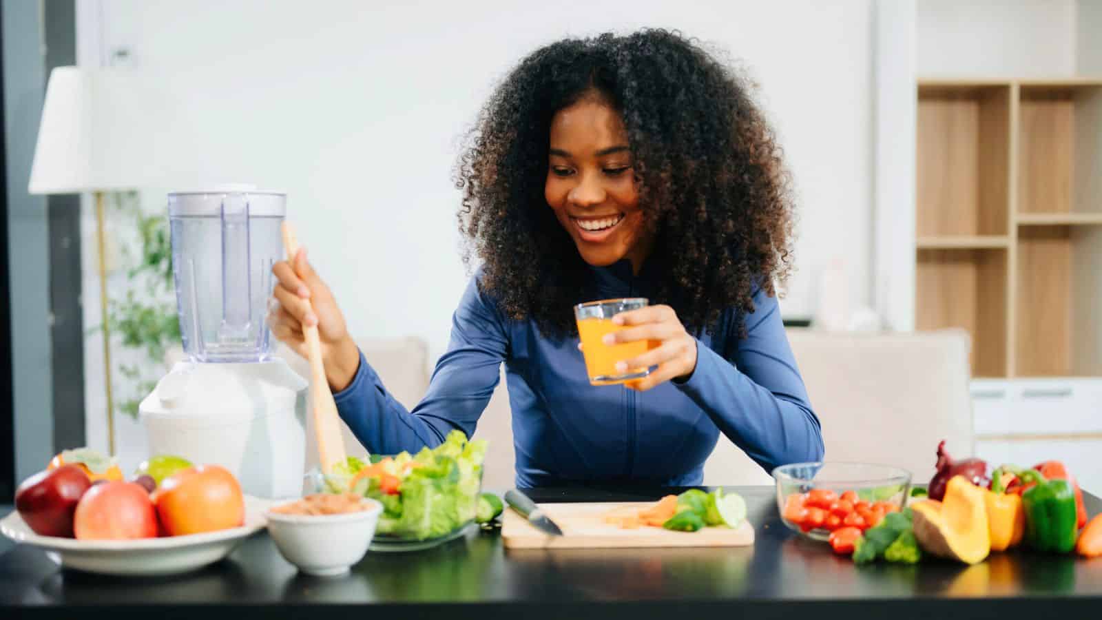 A woman with curly hair sits at a table preparing a meal. She holds a glass of orange juice and uses a wooden spoon. The table is filled with fruits, vegetables, nuts, and a blender. She is smiling while looking at the food.