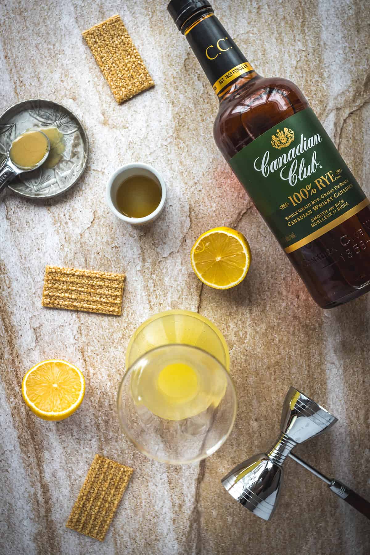 A bottle of Canadian Club 100% Rye whiskey, a jigger, a glass with yellow liquid, sliced lemons, sesame crackers, a spoon with mustard, and a small cup on stone—a perfect setup to craft an enchanting 1001 Arabian Nights cocktail recipe.
