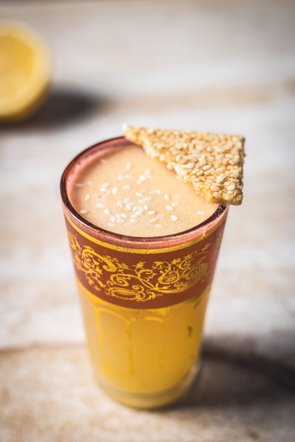 A glass brimming with the 1001 Arabian Nights Cocktail: a frothy yellow drink topped with white sesame seeds and a triangular sesame brittle. The ornate glass features red and gold patterns, with a blurred lemon half in the background.