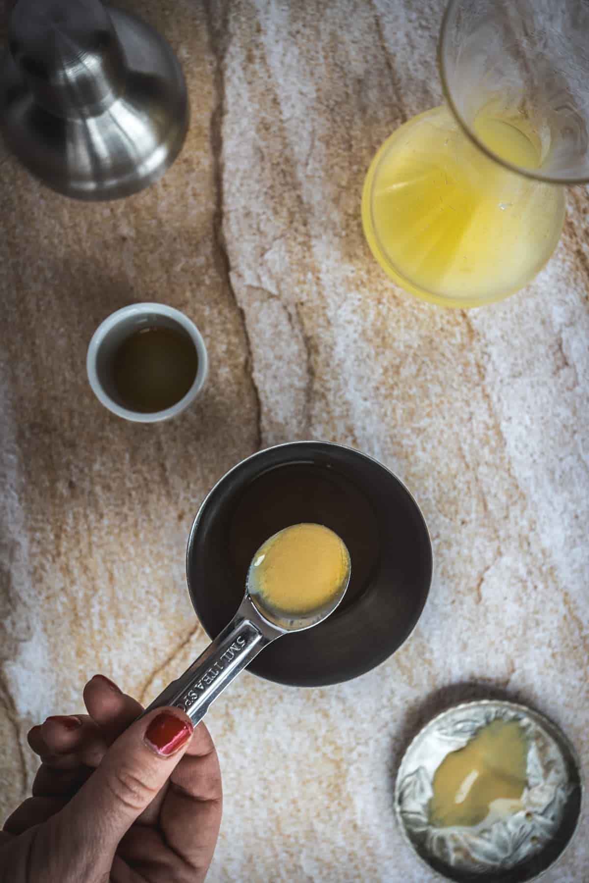 A hand holds a measuring spoon with yellow liquid over a metal cup, preparing the 1001 Arabian Nights Cocktail. Nearby are a carafe with yellow liquid, a small white bowl, and a metal container on a tan marbled surface.