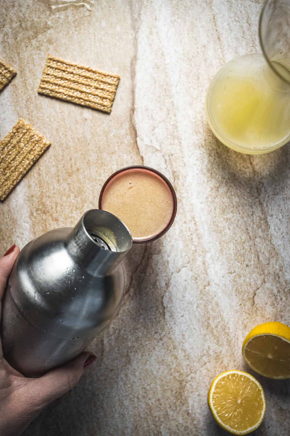 A hand pours a frothy Arabian Nights Cocktail from a shaker into a glass on a stone countertop. Nearby are a glass carafe with yellow liquid, sesame crackers, and a halved lemon—perfect accompaniments for this enchanting cocktail recipe.