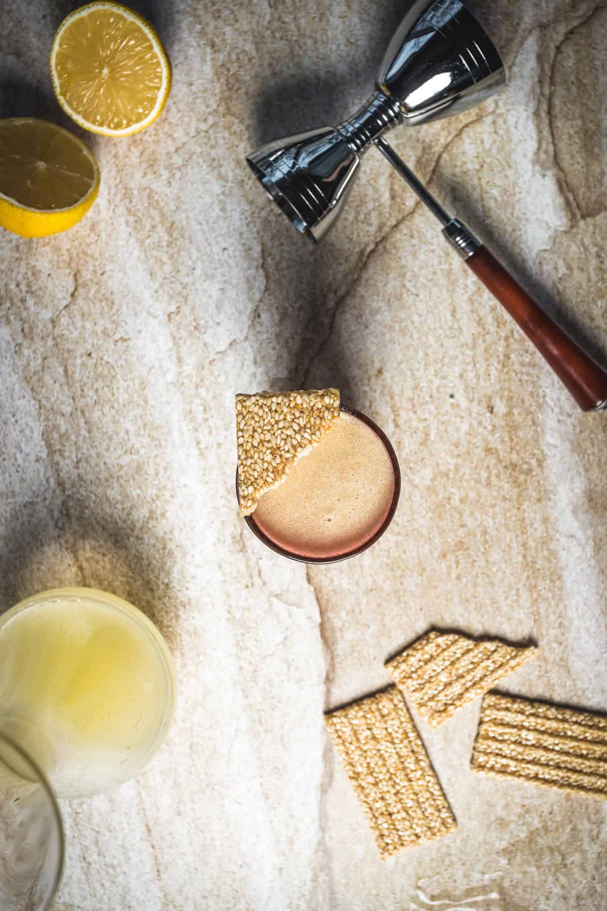A 1001 Arabian Nights Cocktail sits in a glass garnished with sesame brittle on a stone surface. Nearby are lemon halves, a jigger, a glass of yellow liquid, and extra brittle, making this cocktail recipe truly captivating.