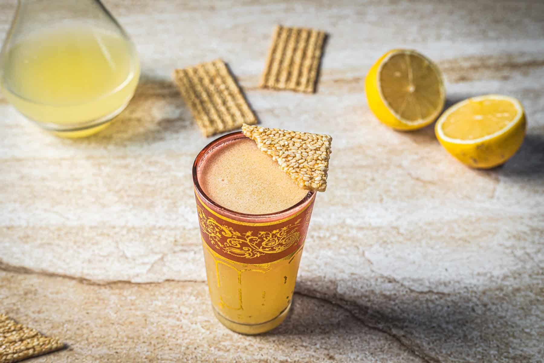 A glass filled with a frothy light brown Arabian Nights Cocktail, garnished with a piece of sesame brittle. In the background, a halved lemon, rectangular crackers, and a glass container with yellow liquid rest on a beige surface.