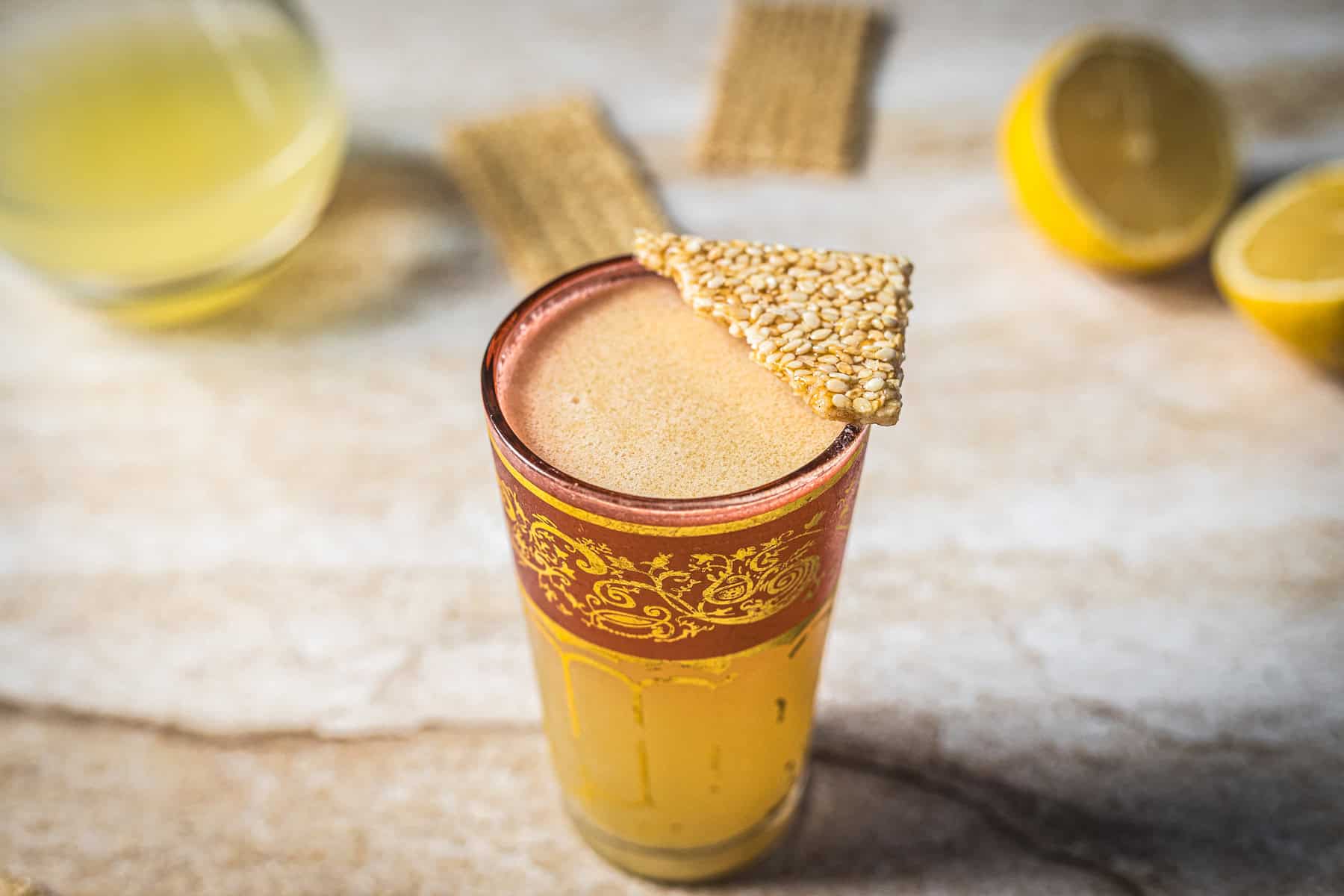A tall glass with an ornate red and gold rim holds a frothy beige Arabian Nights Cocktail, garnished with a piece of sesame brittle. In the background are lemon halves, a small bowl of liquid, and more sesame brittle.