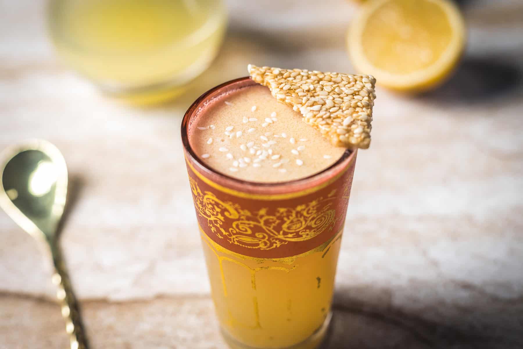 A tall glass with a gold and red decorative rim holds a frothy yellow 1001 Arabian Nights Cocktail, topped with sesame seeds and a piece of sesame brittle. A gold spoon and halved lemon are in the background on a light surface.