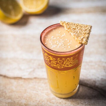 A tall glass with an ornate red and gold rim, filled with a light brown Arabian Nights cocktail topped with sesame seeds and garnished with a triangular sesame crisp. Two halved lemons are blurred in the background.