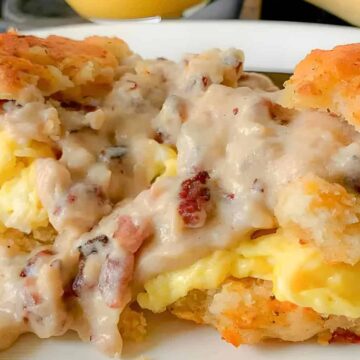 A close-up of a biscuit sandwich filled with scrambled eggs and covered in sausage gravy, served on a white plate. The biscuit is flaky and golden brown, and the gravy contains visible sausage bits.