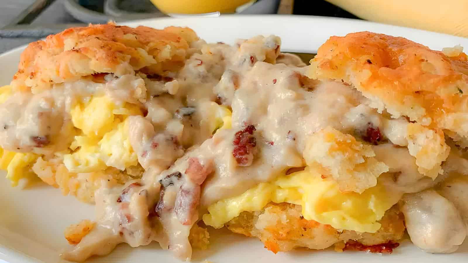 A biscuit sandwich filled with scrambled eggs and covered in thick, creamy gravy with chunks of sausage. In the background, there's part of a stovetop and a yellow item.