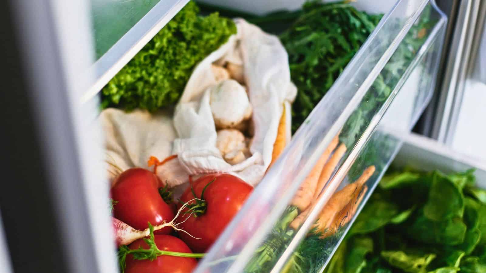 Open refrigerator drawer containing fresh vegetables. There are lettuce, tomatoes, carrots, radishes, and mushrooms. Some items are wrapped in a cloth. The scene is well-lit, highlighting the colors and textures of the produce.