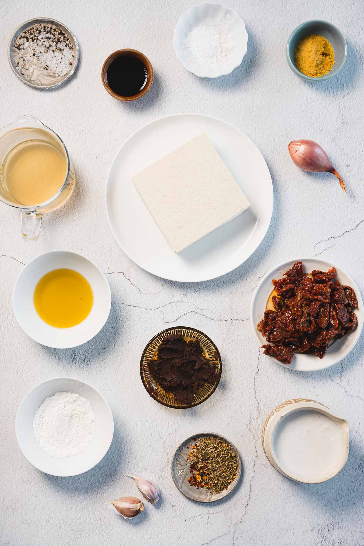 Assorted ingredients are arranged on a light surface, hinting at the preparation of Creamy Sun-Dried Tomato Tofu. A plate with tofu, bowls of sun-dried tomatoes, oil, spices, soy sauce, and garlic cloves surround a shallot and a bowl of liquid for this delicious Marry Me Tofu recipe.