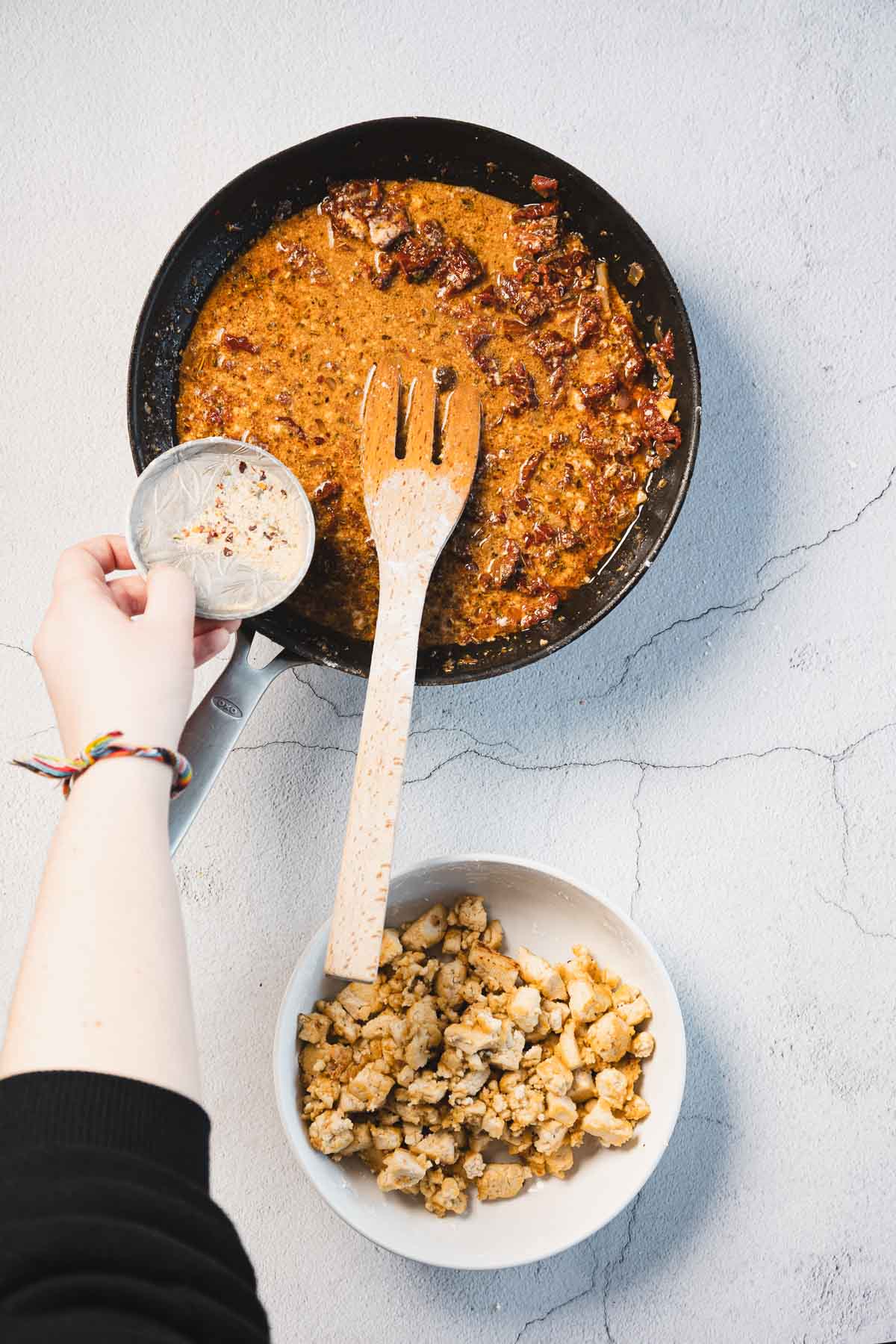 A person adds a white powdery ingredient into a black skillet containing a creamy sun-dried tomato tofu sauce. A wooden spatula rests in the skillet, and a white bowl with cooked cauliflower lies nearby on the textured surface.