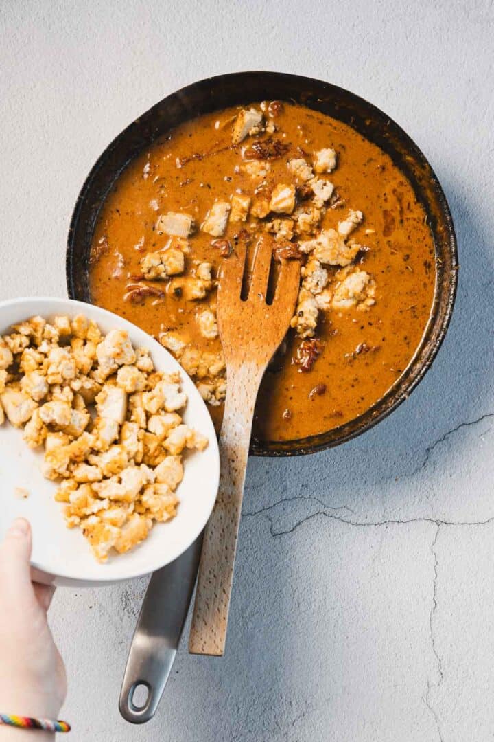 A frying pan with chunks of chicken in a rich, brown sauce reminiscent of a creamy sun-dried tomato tofu dish. A wooden spatula rests nearby while a hand holds a white plate above, ready to serve more savory delights on the textured light gray surface.