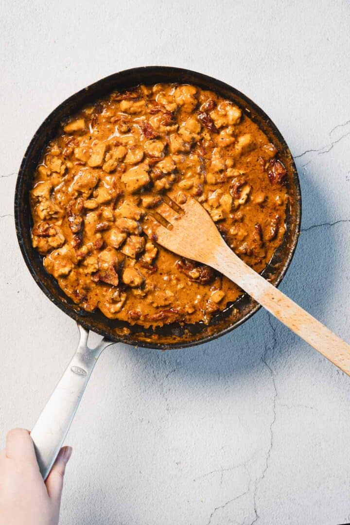 A skillet brims with creamy, browned chicken pieces in a rich sun-dried tomato sauce, sitting on a light gray textured surface. A wooden spatula rests inside, and a hand holds the handle. This delightful fusion could very well be called "Marry Me Tofu.