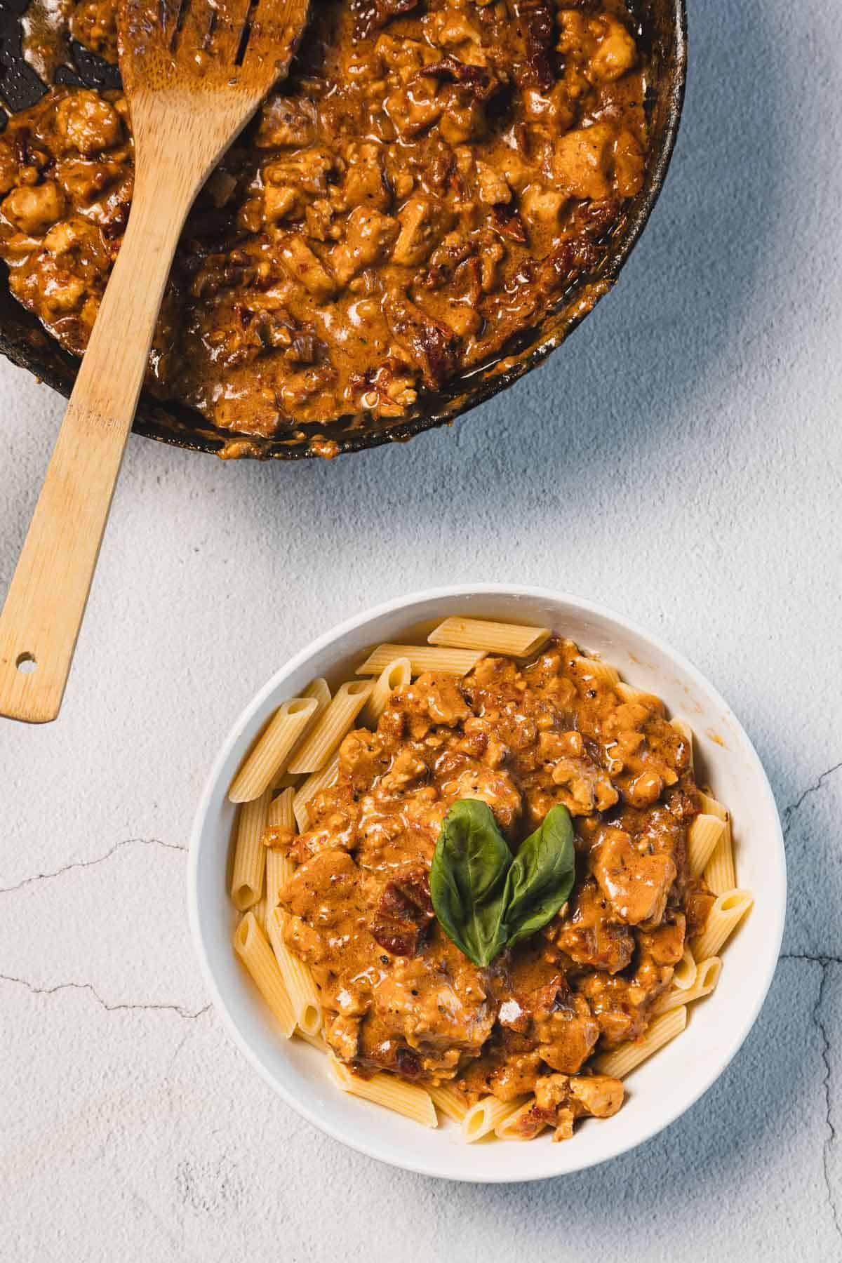 A bowl of penne pasta topped with a creamy sun-dried tomato tofu sauce garnished with a basil leaf. A wooden spoon rests in a pan filled with the same marry me tofu sauce beside the bowl, all set on a light stone surface.