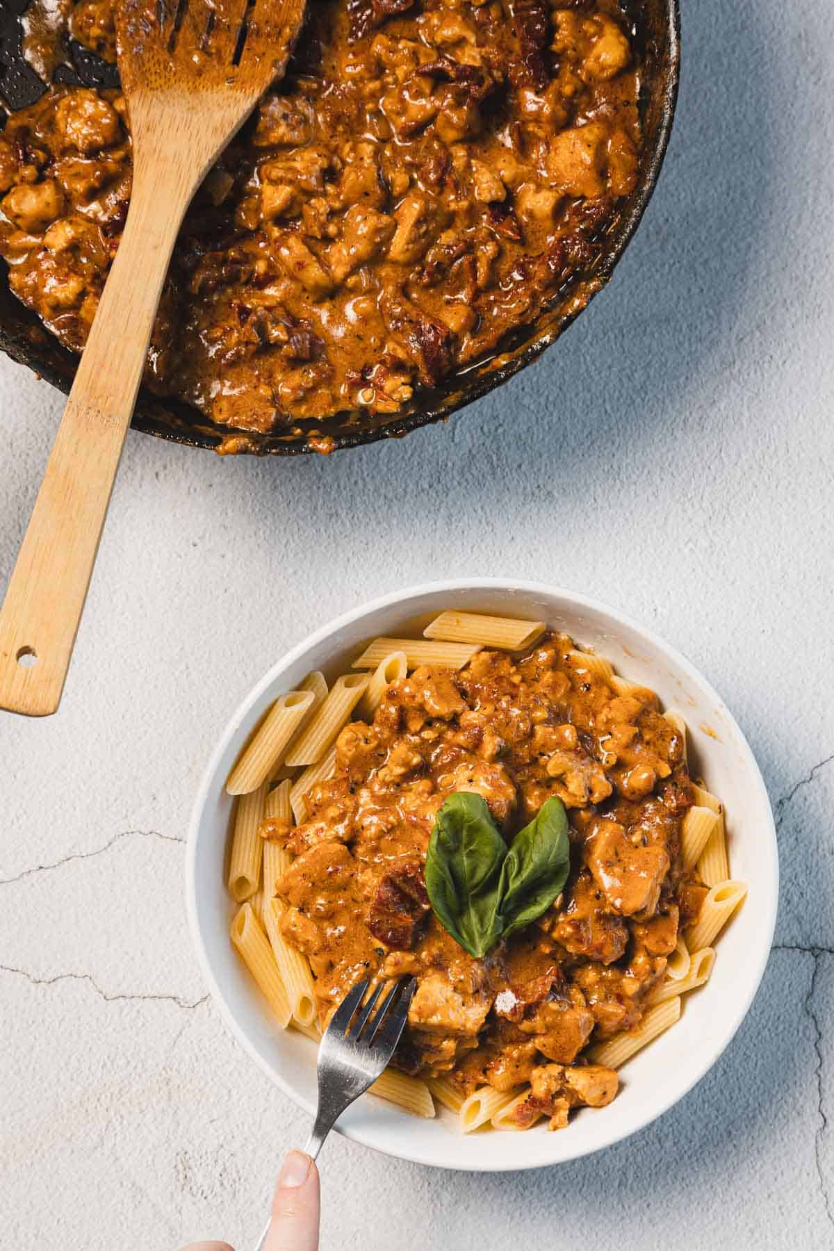 A bowl of penne pasta topped with a chunky tomato-based sauce and garnished with fresh basil. Nearby, a skillet filled with creamy sun-dried tomato tofu sauce beckons, ready to marry flavors together. A fork rests beside the bowl on the light textured surface.