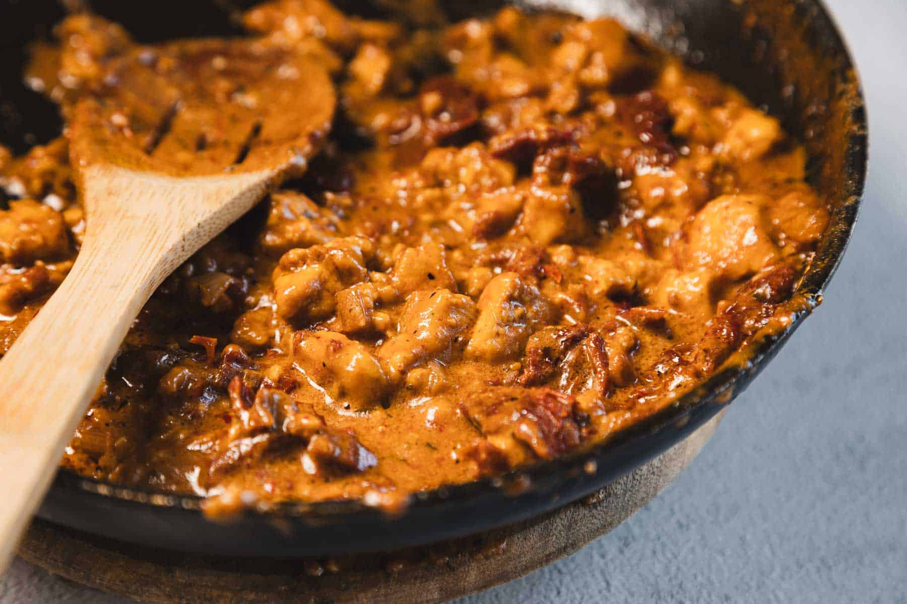A close-up of a frying pan cradles a rich, brown stew with chunks of meat and visible herbs. A wooden spoon rests beside creamy sun-dried tomato tofu, lovingly dubbed "Marry Me Tofu," infusing the dish with depth. The surface beneath the pan is light gray.