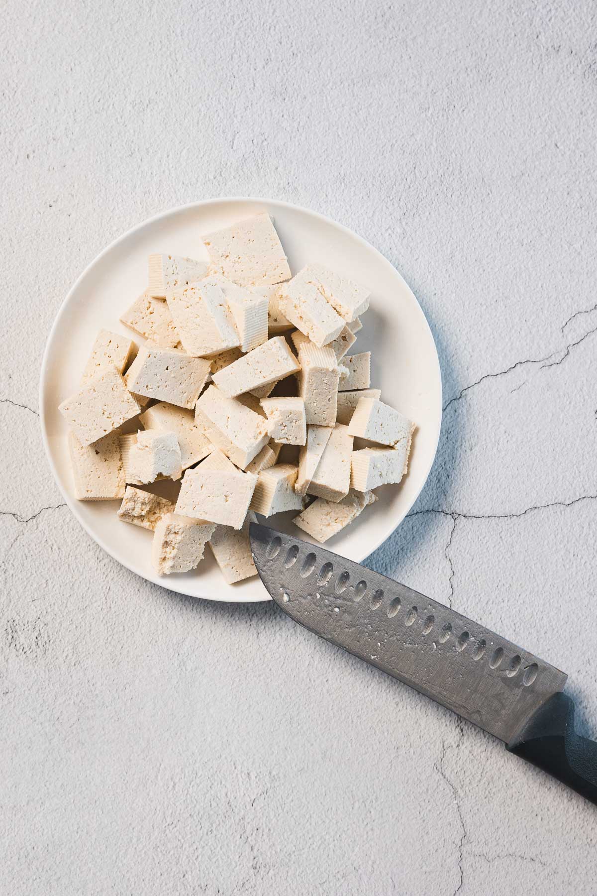 A white plate filled with cubed Creamy Sun-Dried Tomato Tofu, known as Marry Me Tofu, sits on a cracked white surface. A large chef's knife rests beside the plate, partially under some tofu cubes. The setting exudes a minimalist and clean look.