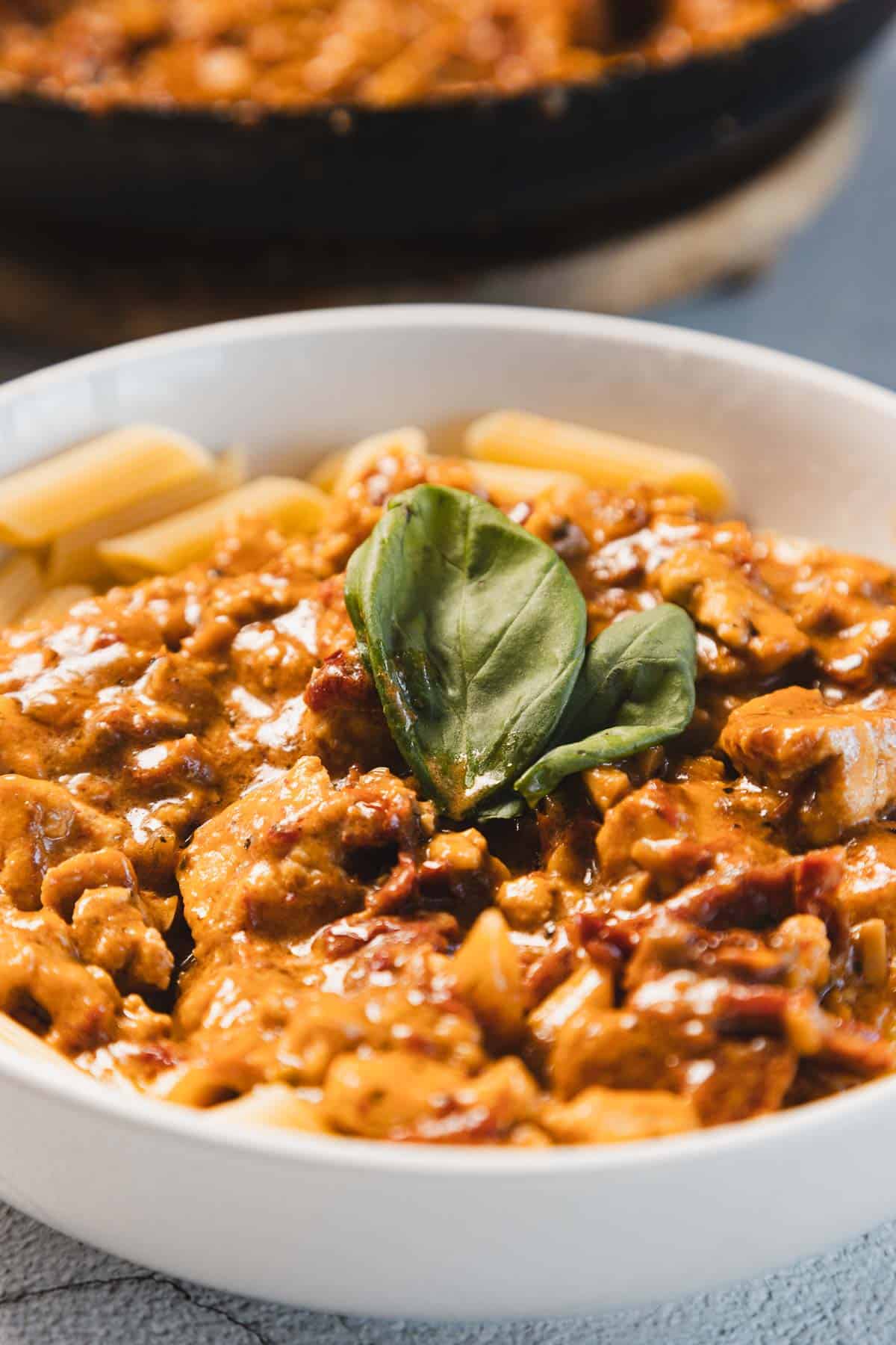 A white bowl filled with penne pasta topped with a creamy sun-dried tomato sauce and pieces of chicken. A fresh basil leaf is placed on top as garnish, while a blurred skillet lingers in the background, hinting at the secrets of Marry Me Tofu.
