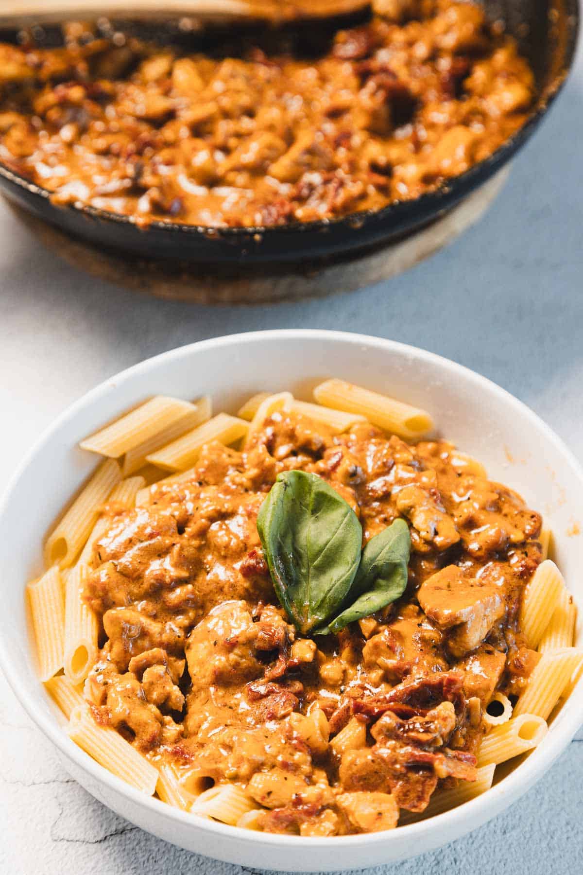 A white bowl filled with penne pasta topped with a luscious creamy sun-dried tomato sauce, garnished with a basil leaf. In the background, a pan brims with more of the "Marry Me Tofu" sauce, promising irresistible flavor in every bite.