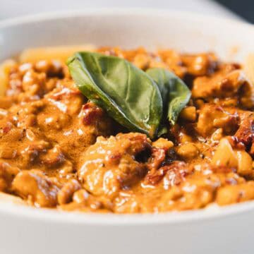 A bowl of rigatoni pasta topped with a creamy sun-dried tomato sauce featuring chunks of tofu and garnished with a fresh basil leaf. The dish, known as "Marry Me Tofu," is elegantly presented in a white bowl.