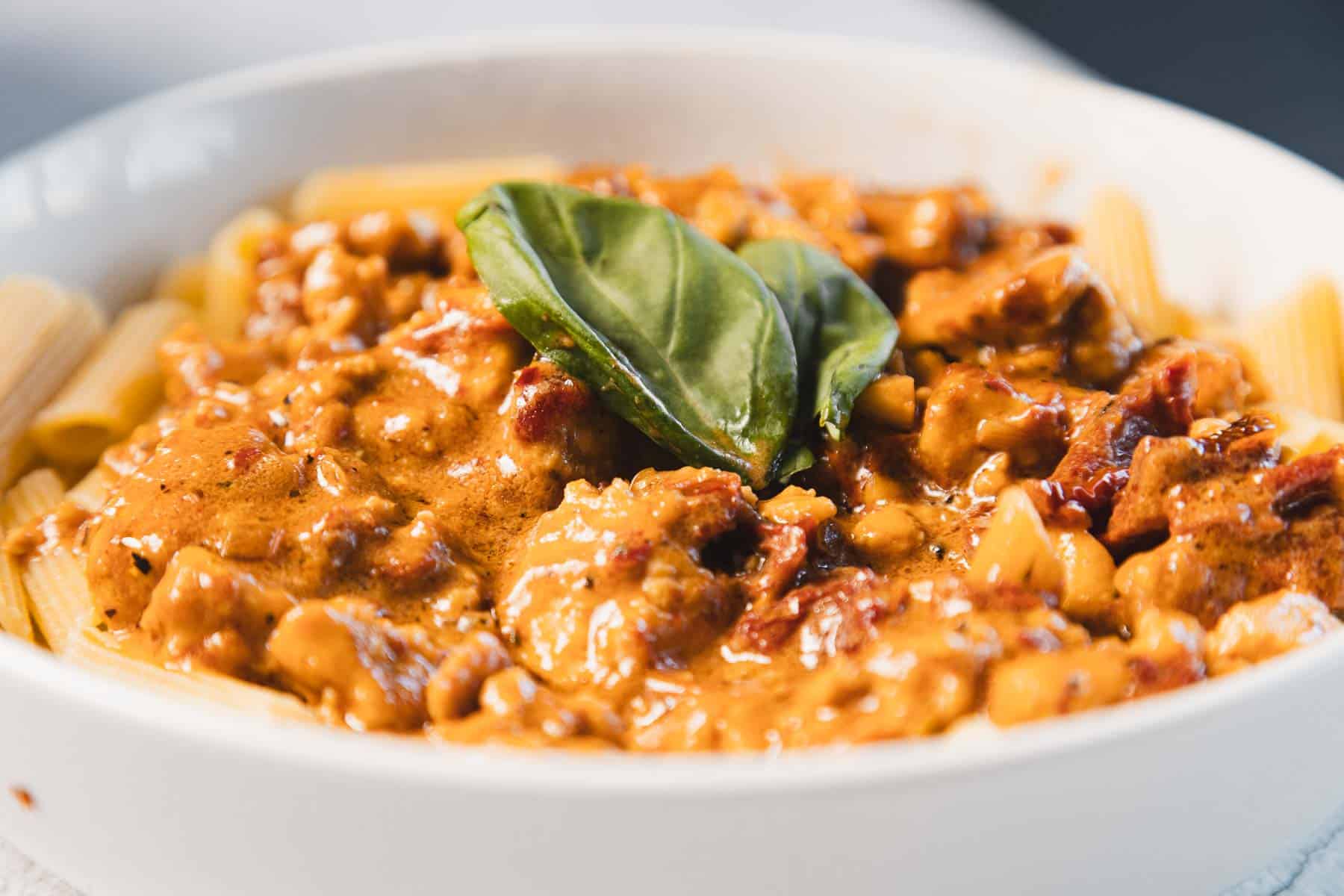 A bowl of rigatoni pasta topped with a creamy sun-dried tomato sauce featuring chunks of tofu and garnished with a fresh basil leaf. The dish, known as "Marry Me Tofu," is elegantly presented in a white bowl.