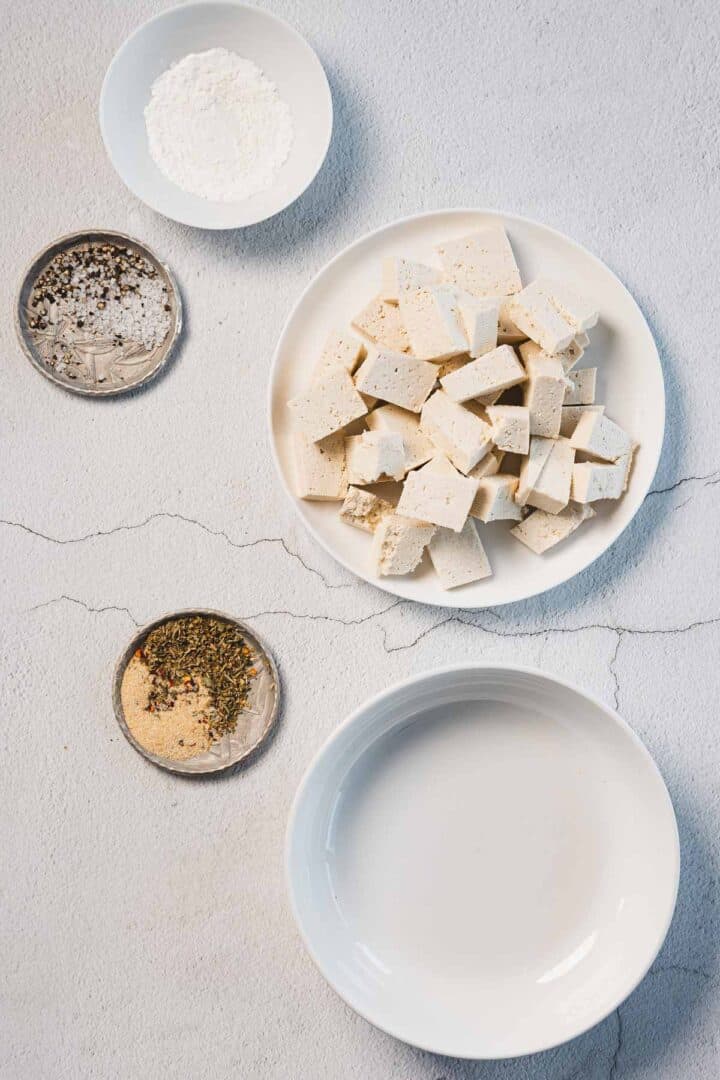 A white plate featuring creamy sun-dried tomato tofu, also known as Marry Me Tofu, rests on a textured surface. Nearby are three small containers: one with white powder, another with beige powder and herbs, and a third with crushed black and white specks. An empty white bowl is also present.