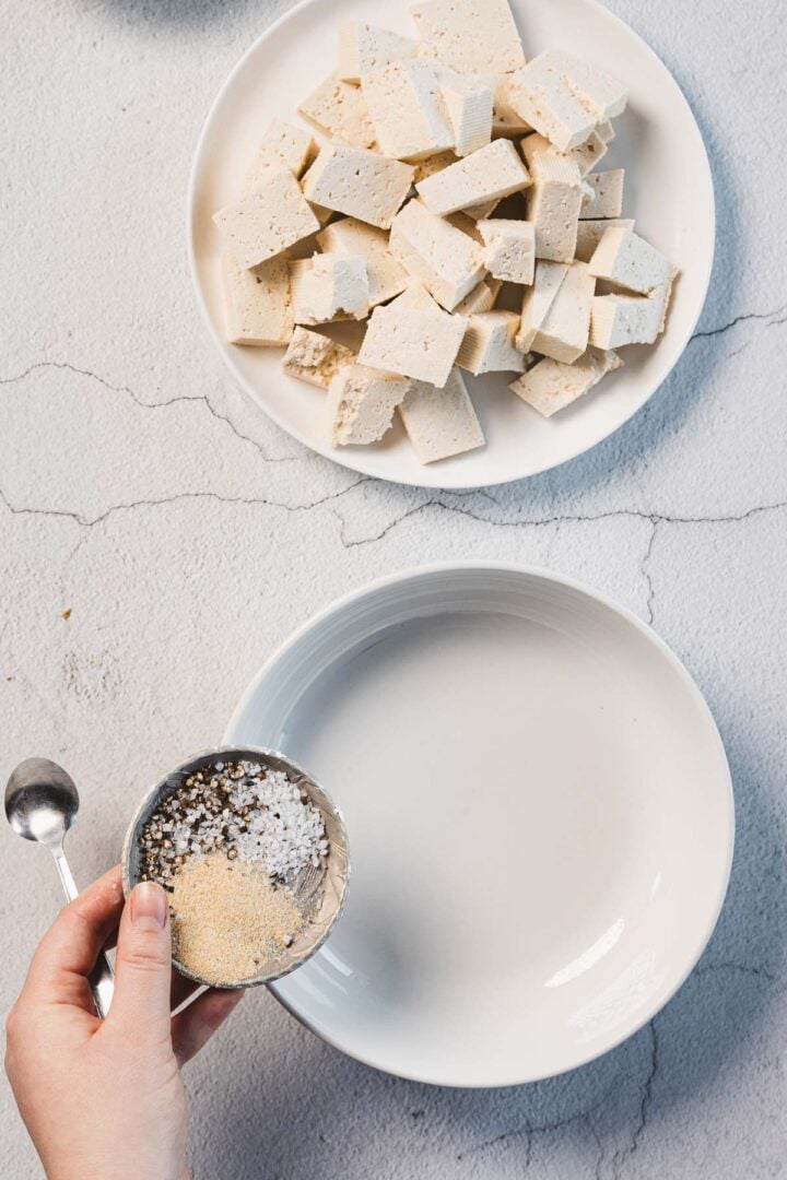 A hand holds a small bowl of salt and pepper above a larger empty white plate, ready for its next creation. Nearby, on a cracked white surface, rests cubed tofu destined to become Creamy Sun-Dried Tomato Tofu. A teaspoon stands by, eager to assist in the Marry Me Tofu endeavor.