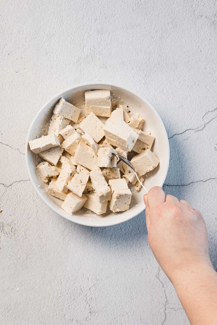 A hand holding a fork is reaching into a white bowl filled with cubed tofu, glistening with savory creamy sun-dried tomato seasoning, aptly named Marry Me Tofu. The bowl rests on a textured, light-colored surface.