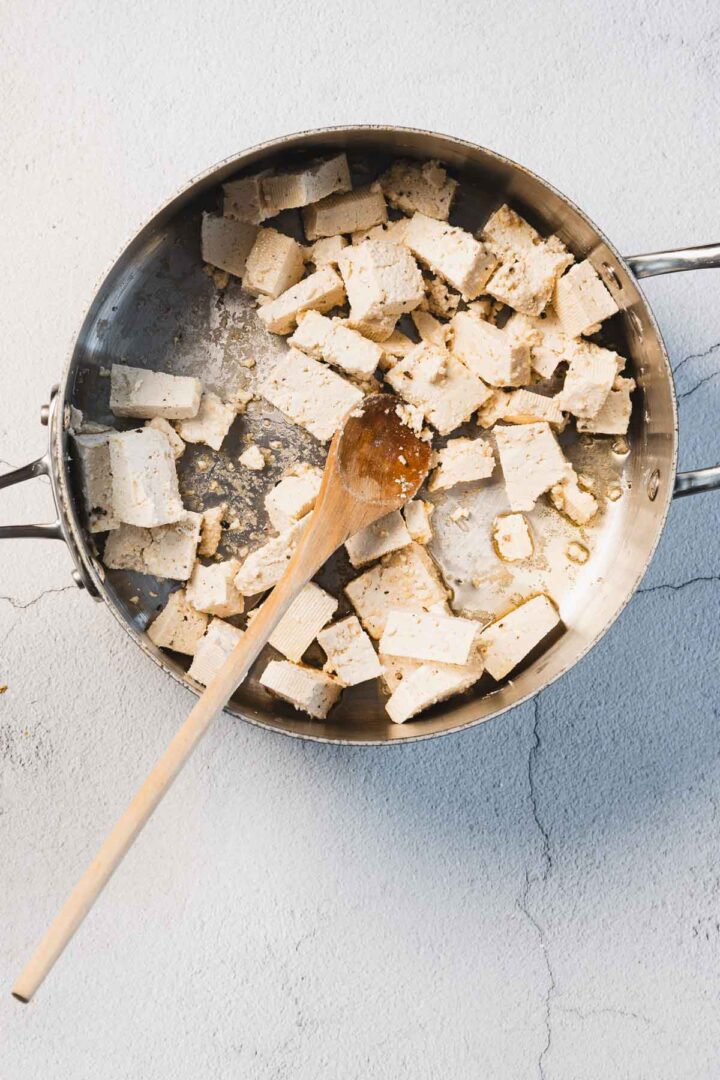 A metal pan with cubed tofu pieces, some slightly browned and kissed by creamy sun-dried tomato goodness, rests on a light, textured surface. A wooden spoon lies inside, hinting at the delicious "Marry Me Tofu" dish yet to be enjoyed.