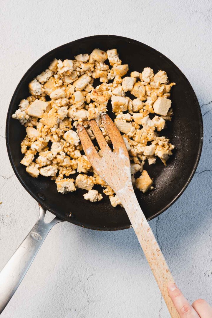 A frying pan containing Creamy Sun-Dried Tomato Tofu, also known as Marry Me Tofu, sits on a light surface. A person's hand holds a wooden spatula, stirring the tofu pieces, which are golden brown and slightly crumbly in texture.