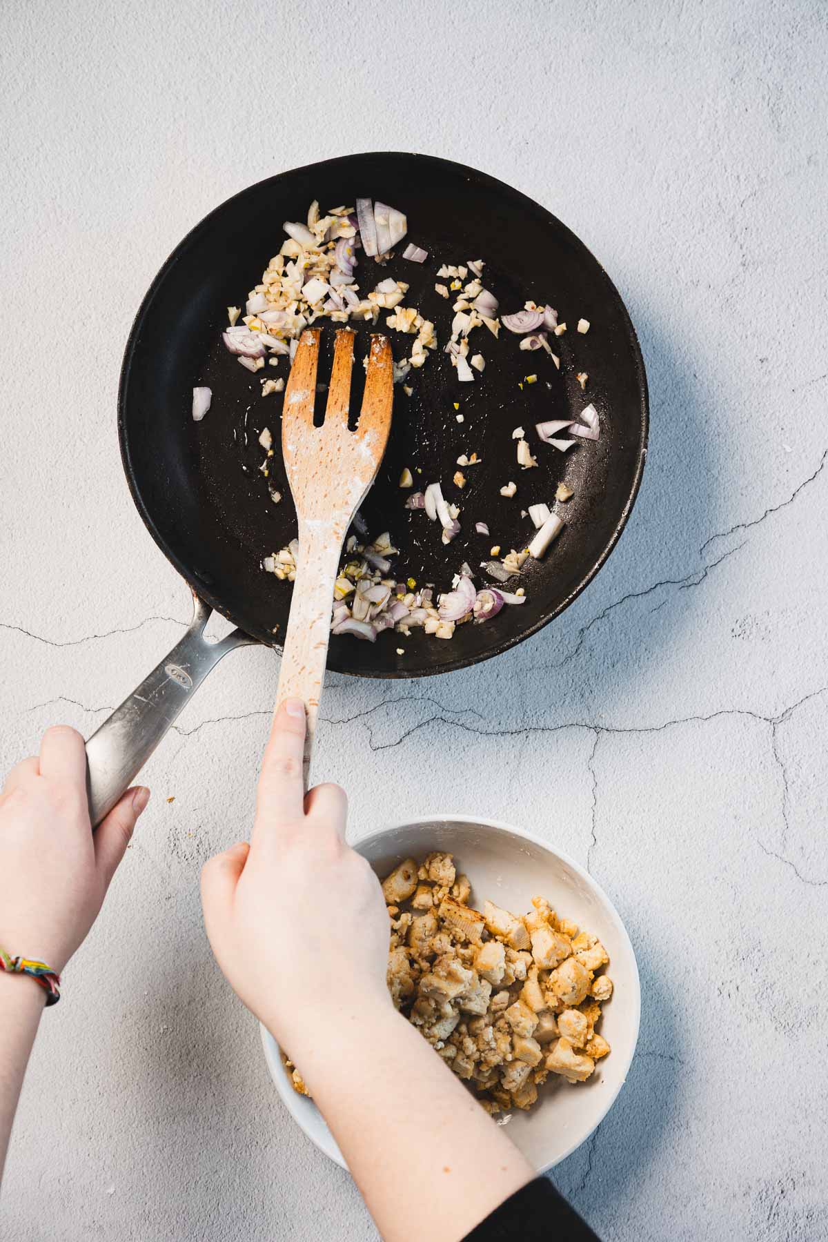 Hands are shown using a wooden spatula to cook chopped onions and garlic in a black frying pan, preparing the base for a creamy sun-dried tomato tofu dish. Below the pan, there is a white bowl containing crumbled tofu. The background is a light-colored, textured surface.