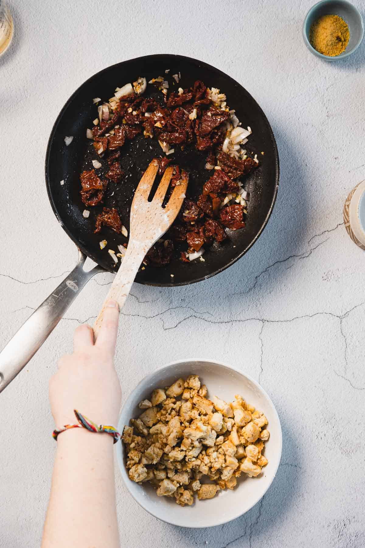 A top view of someone stirring diced sundried tomatoes and onions in a black frying pan with a wooden spatula, crafting the perfect base for Creamy Sun-Dried Tomato Tofu. On the side, a bowl of crumbled tofu or cooked chicken rests on a light gray textured surface.