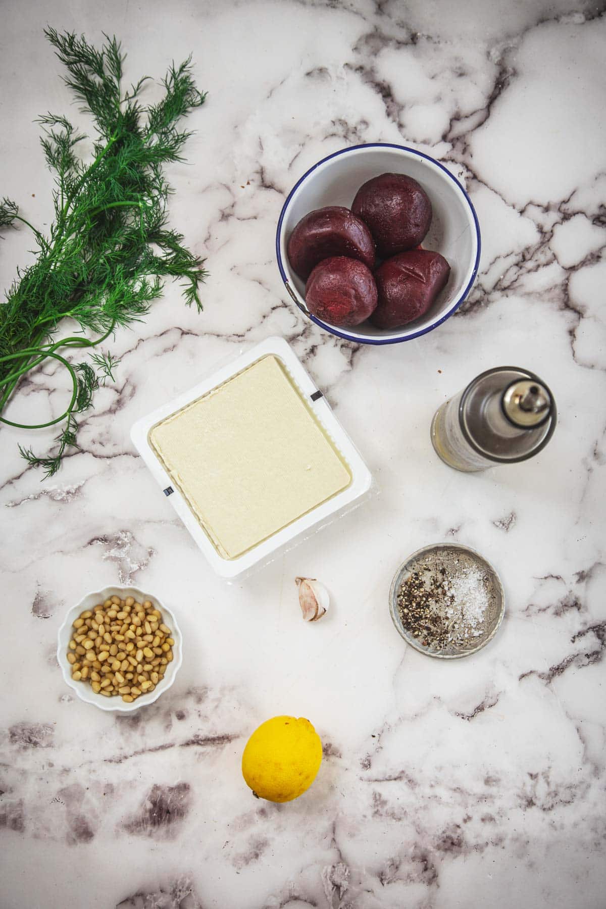 A marble surface with a block of tofu, a bunch of fresh dill, a bowl of cooked beets, a bottle of olive oil, pine nuts, garlic, lemon, and a dish of salt and pepper—perfect for crafting vibrant beet and tofu toast.