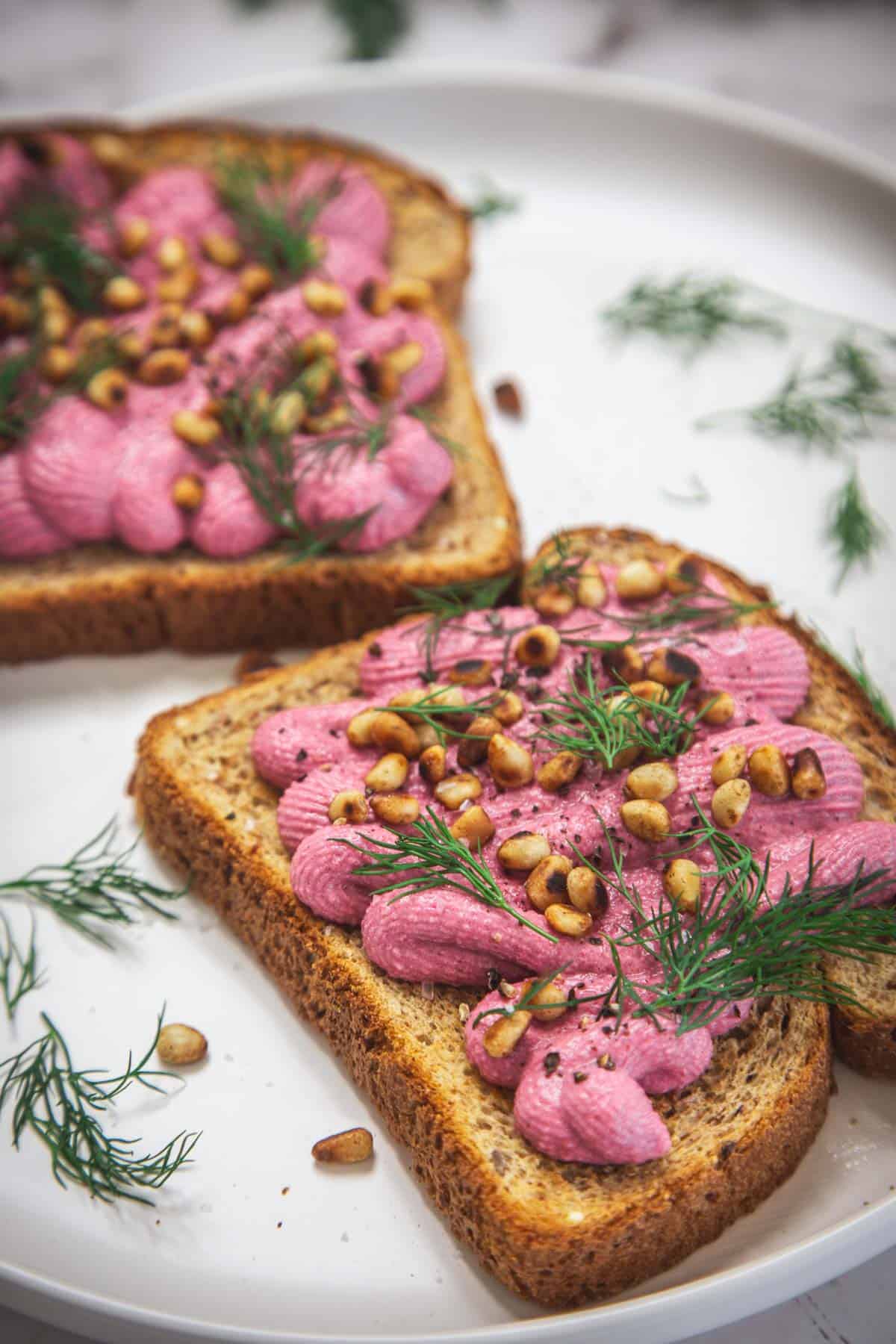 Two slices of toasted bread topped with creamy beet spread, garnished with fresh dill and toasted pine nuts, served on a white plate with extra dill scattered around.