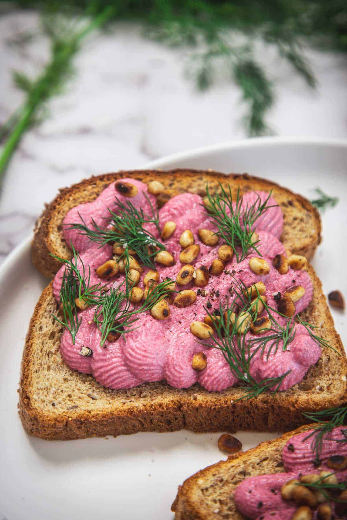 Two slices of toast on a white plate topped with vibrant pink beet hummus, toasted pine nuts, and fresh dill sprigs, set against a backdrop of blurred green herbs.