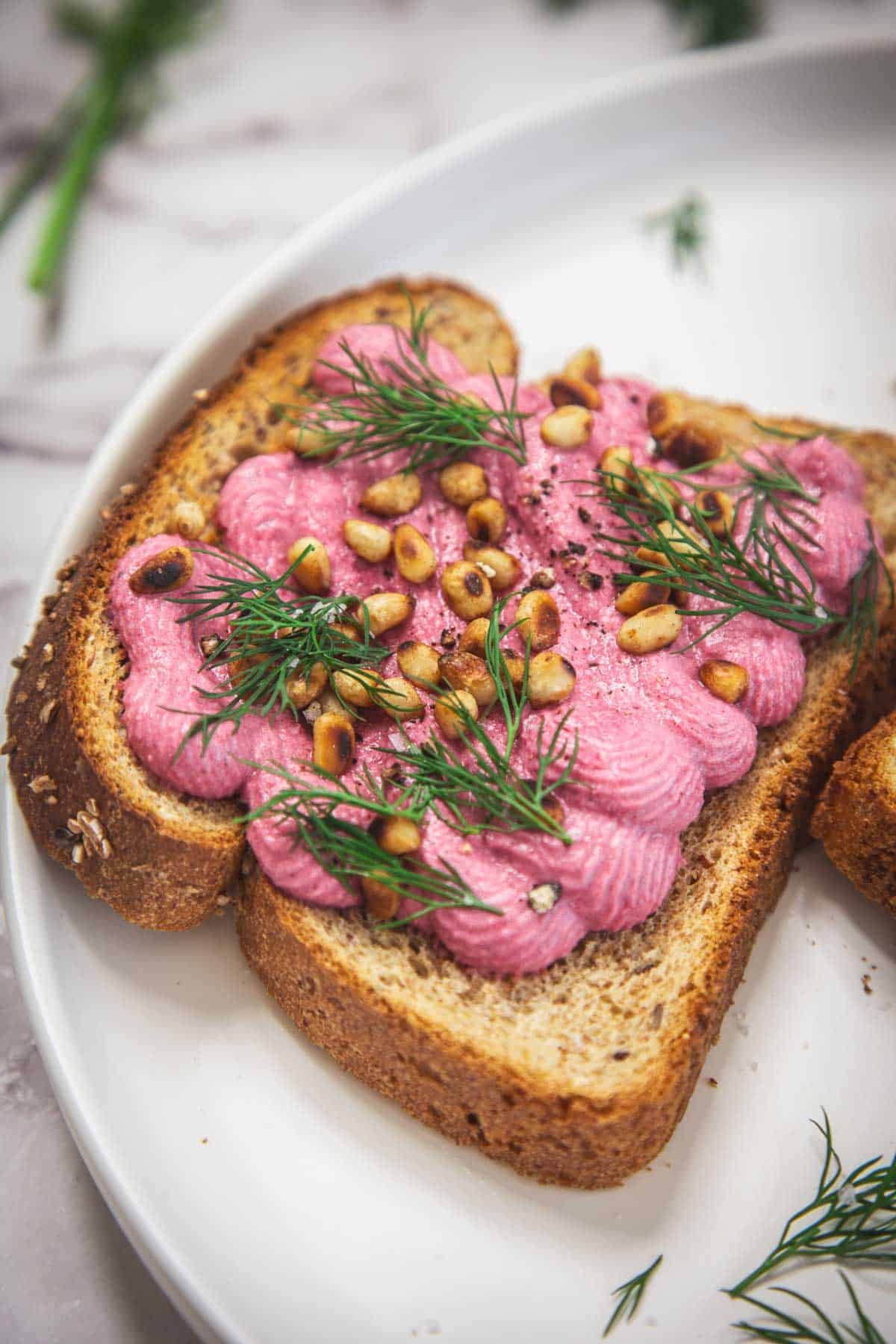 A slice of whole grain toast topped with a creamy beet and tofu spread, garnished with fresh dill and toasted pine nuts, served on a white plate.