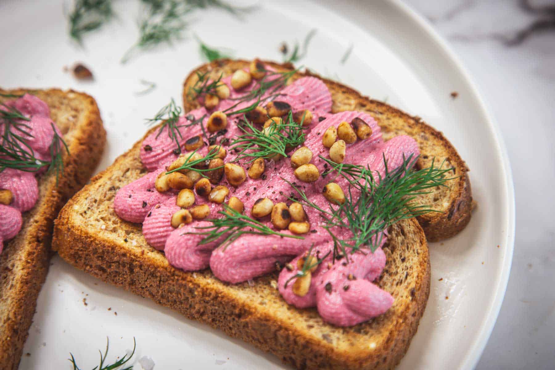 A slice of whole grain toast topped with a vibrant beet and tofu spread, sprinkled with pine nuts and fresh dill, served on a white plate. Another slice of similarly topped toast is partially visible to the side.