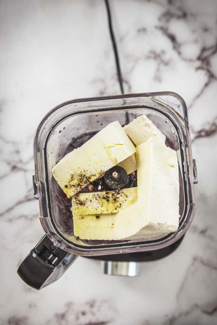 A top-down view of a blender containing large rectangular pieces of tofu, ground black pepper, and yellow seasoning on a marble countertop—perfect for making a creamy spread for toast.
