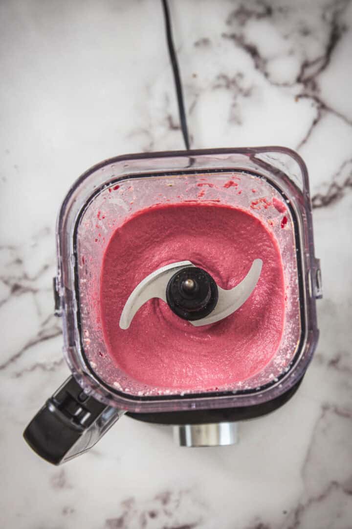 A top-down view of a blender containing a smooth, bright pink beet mixture. The blender is placed on a white and gray marbled surface, with its metal blade visible in the center of the vibrant blend.