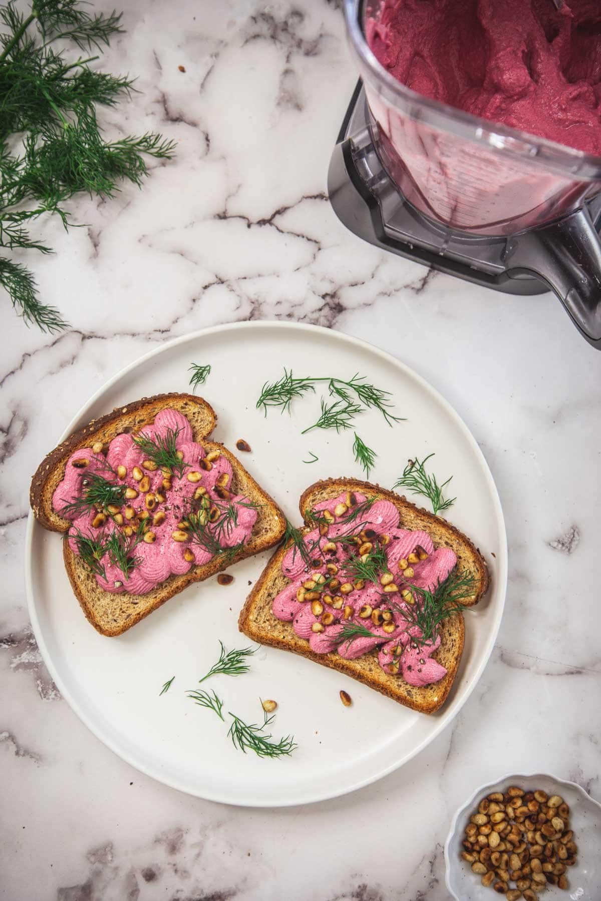 Two slices of toast on a white plate topped with creamy beet spread, pine nuts, and fresh dill. A blender with beet-tofu spread, sprigs of dill, and a small bowl of pine nuts sit nearby on a marble countertop.