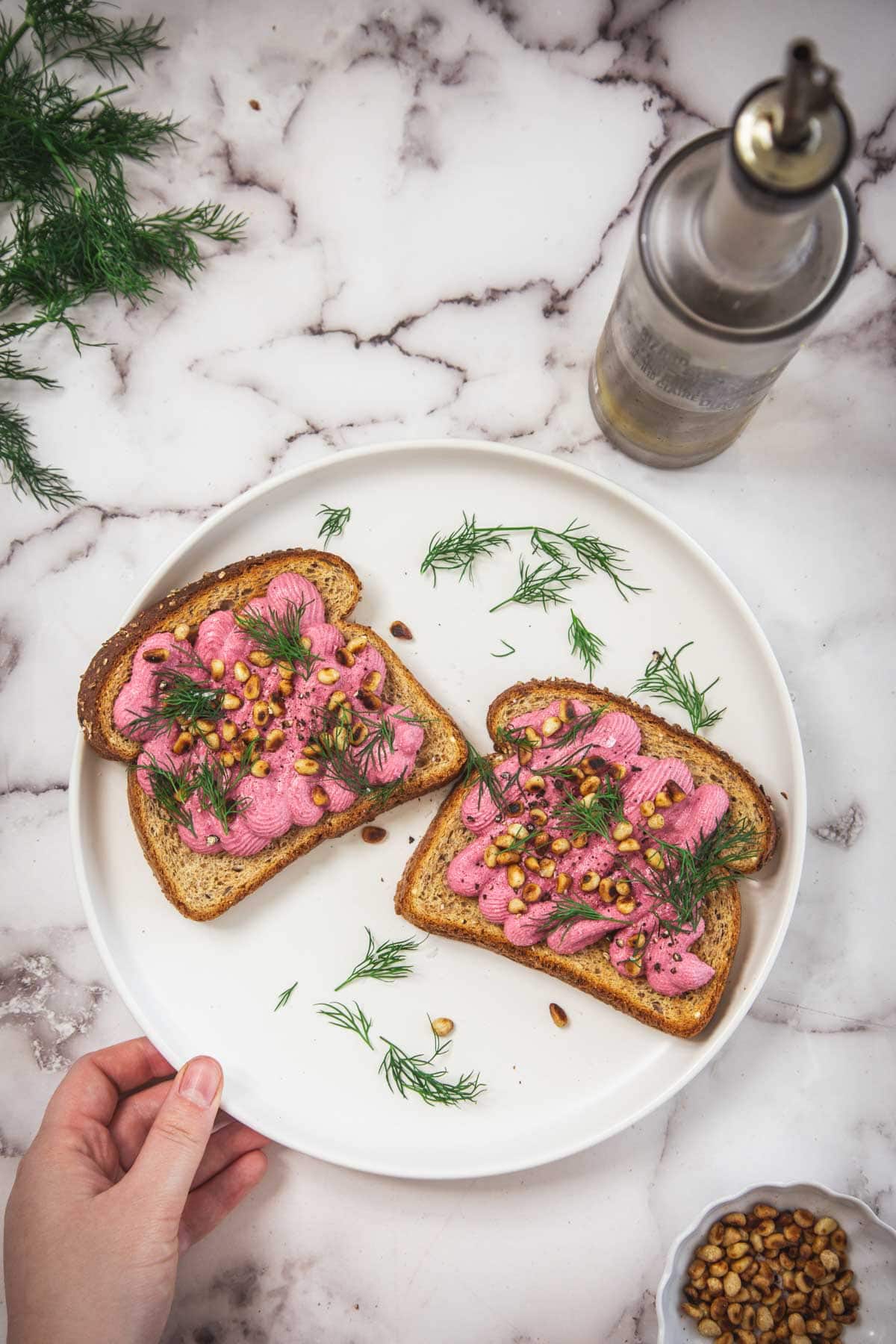 A hand holds a white plate with two slices of beet toast topped with pink spread, pine nuts, and dill. Fresh dill, a bottle of oil, and a small bowl of pine nuts are on a marble countertop.
