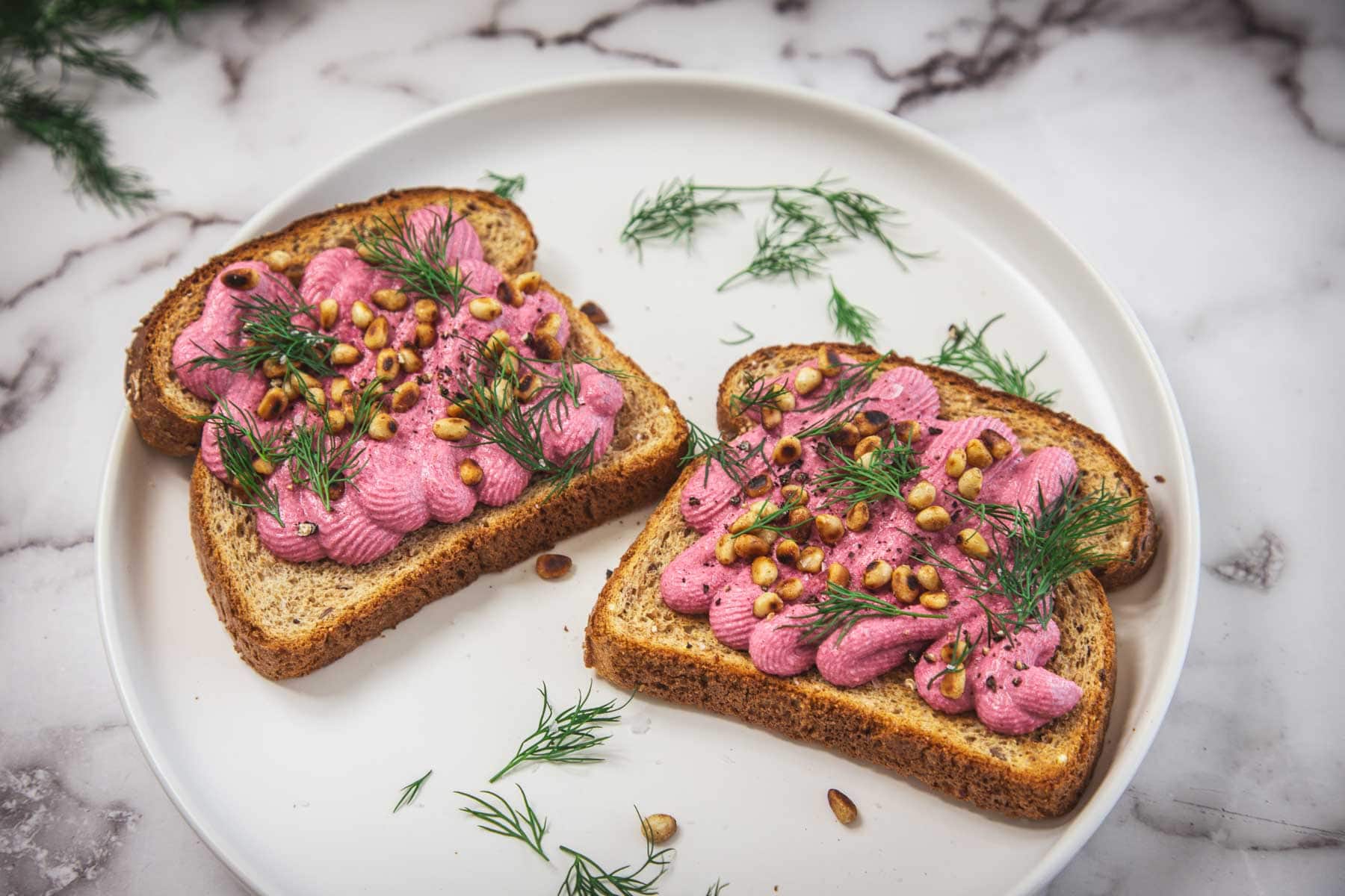 Two slices of whole grain toast topped with a pink beet spread, garnished with fresh dill and pine nuts, are placed on a white plate with sprigs of dill scattered around.