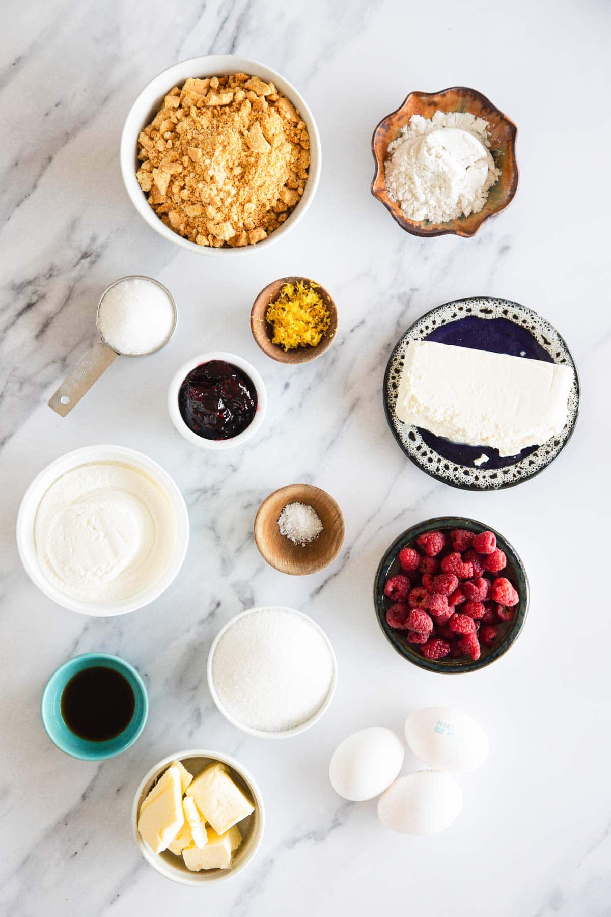 Overhead view of baking ingredients for Raspberry Ricotta Cheesecake on a marble surface—eggs, raspberries, butter, ricotta, cream cheese, sugar, flour, crushed cookies, vanilla, jam, lemon zest, and salt in bowls and dishes.