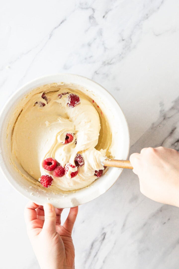 A person mixes a creamy batter for Raspberry Ricotta Cheesecake with fresh raspberries in a white bowl using a wooden spatula on a marble countertop.