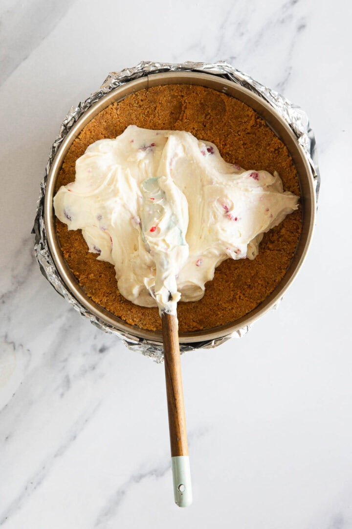 A springform pan lined with foil contains a graham cracker crust, with a spatula spreading a thick layer of Raspberry Ricotta Cheesecake filling—rich and creamy, studded with fruit pieces—on top.
