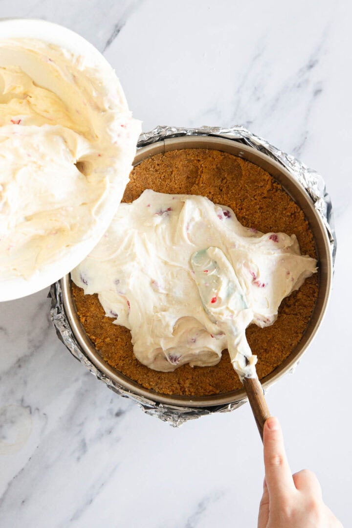 A hand spreads creamy Raspberry Ricotta Cheesecake filling with pieces of fruit over a graham cracker crust in a foil-lined springform pan. Above the pan, a bowl with more cheesecake filling rests on a white marble surface.