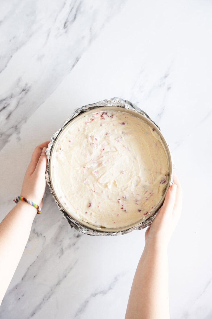 A person holds a round, foil-wrapped cake pan filled with unbaked Raspberry Ricotta Cheesecake batter with small red specks on a white marble surface. The person wears a rainbow bracelet on one wrist.